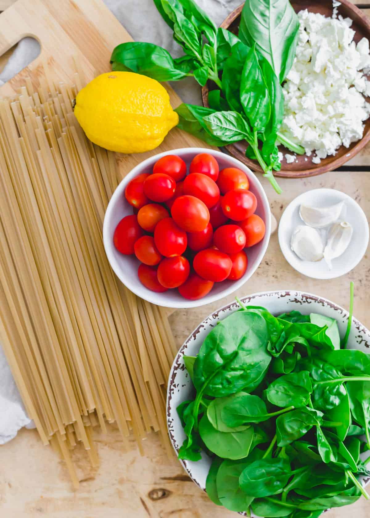 Uncooked linguine, cherry tomatoes, garlic, feta, basil, lemon and baby spinach on a wood surface.