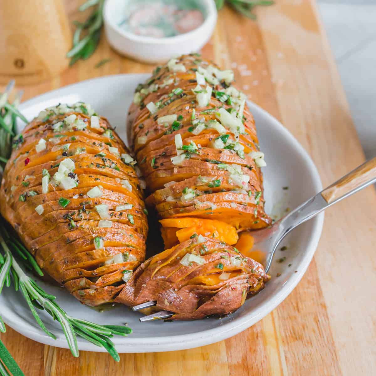 A forkful of roasted hasselback sweet potatoes on a plate with fresh rosemary sprigs.