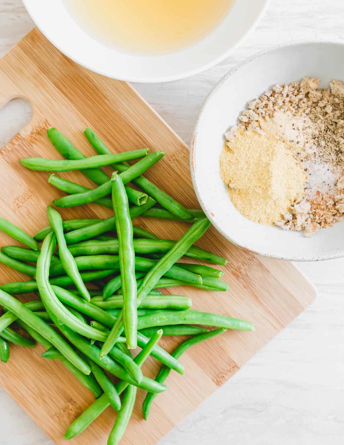 Fresh green beans on a cutting board with breadcrumb mixture in a bowl to the side and aquafaba in another bowl.