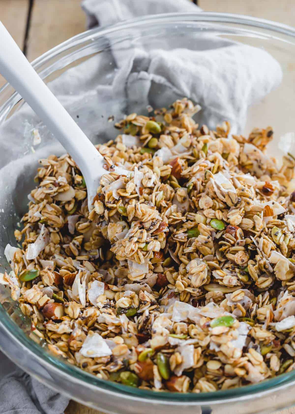 Oats, almonds, coconut, pepitas and chia seeds tossed with wet ingredients to make granola in a glass bowl.
