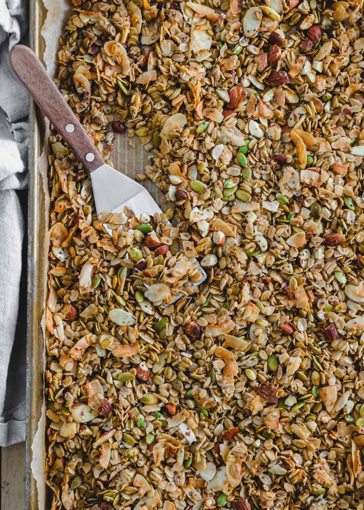 Oat coconut granola with almonds spread out on a baking sheet with a spatula.