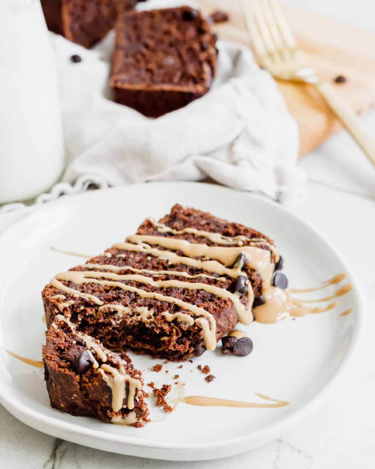 A slice of healthier chocolate banana bread on a plate drizzled in peanut butter with chocolate chips scattered around it.