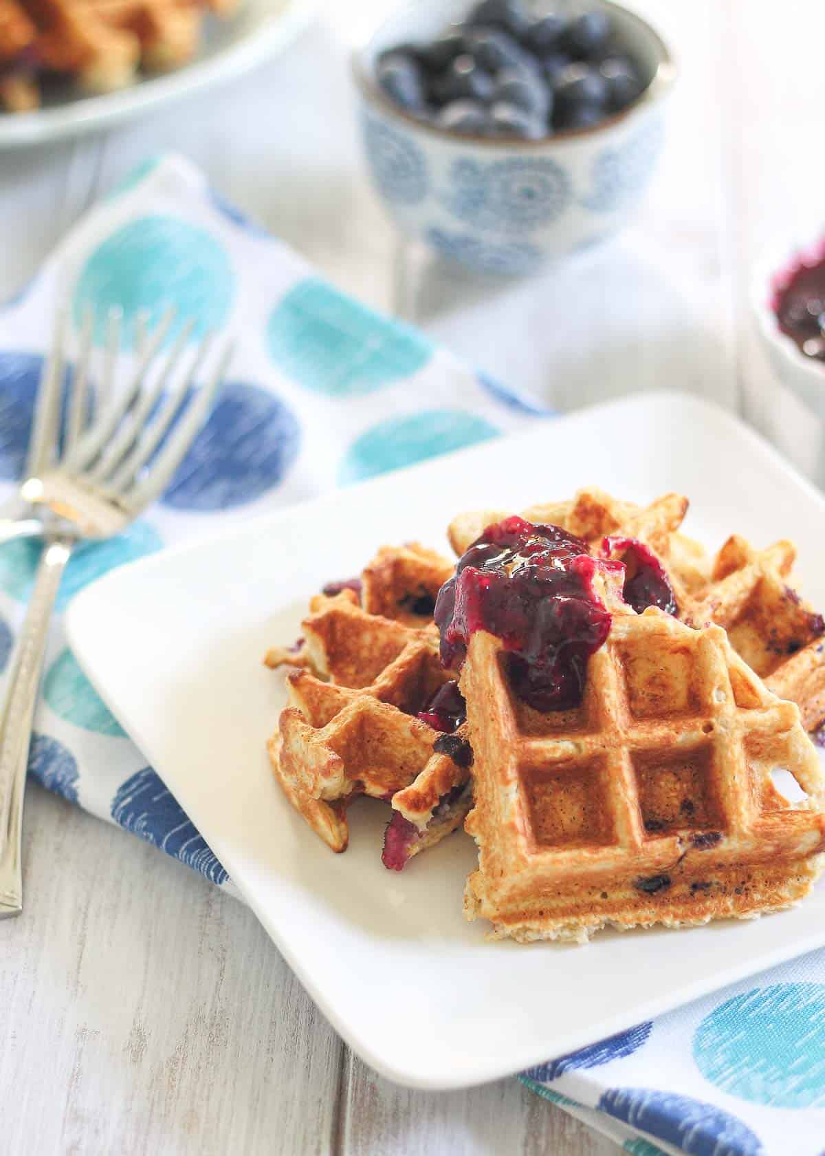 Pieces of oatmeal waffles on a white plate topped with blueberry fruit sauce.