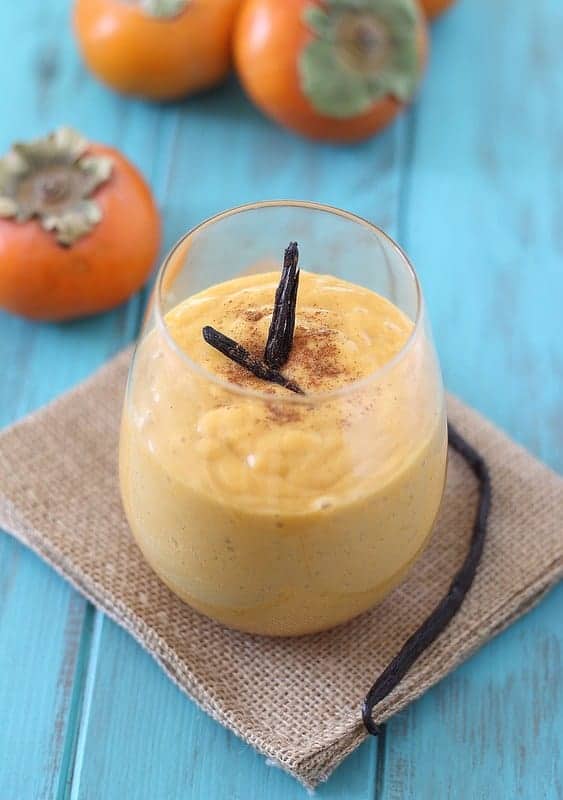 A persimmon smoothie in a stemless glass topped with garnish next to whole persimmons.