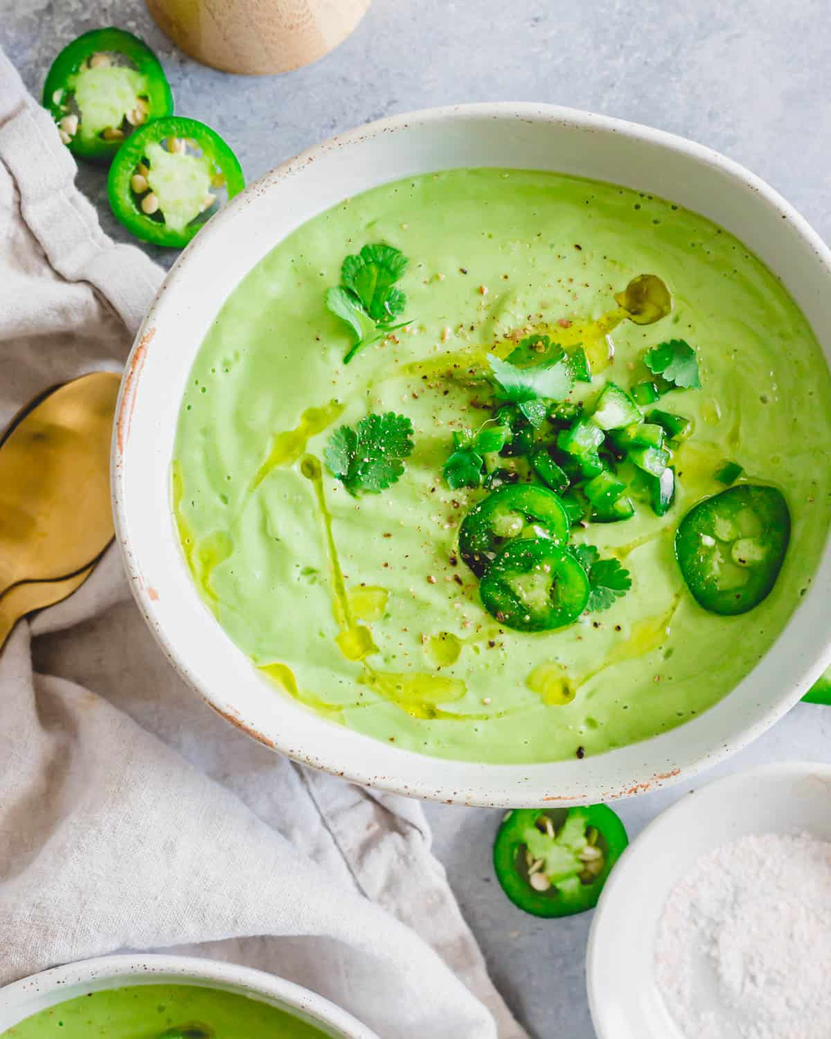 Chilled avocado soup in a bowl drizzled with olive oil and garnished with jalapeño peppers, cilantro, salt and pepper.