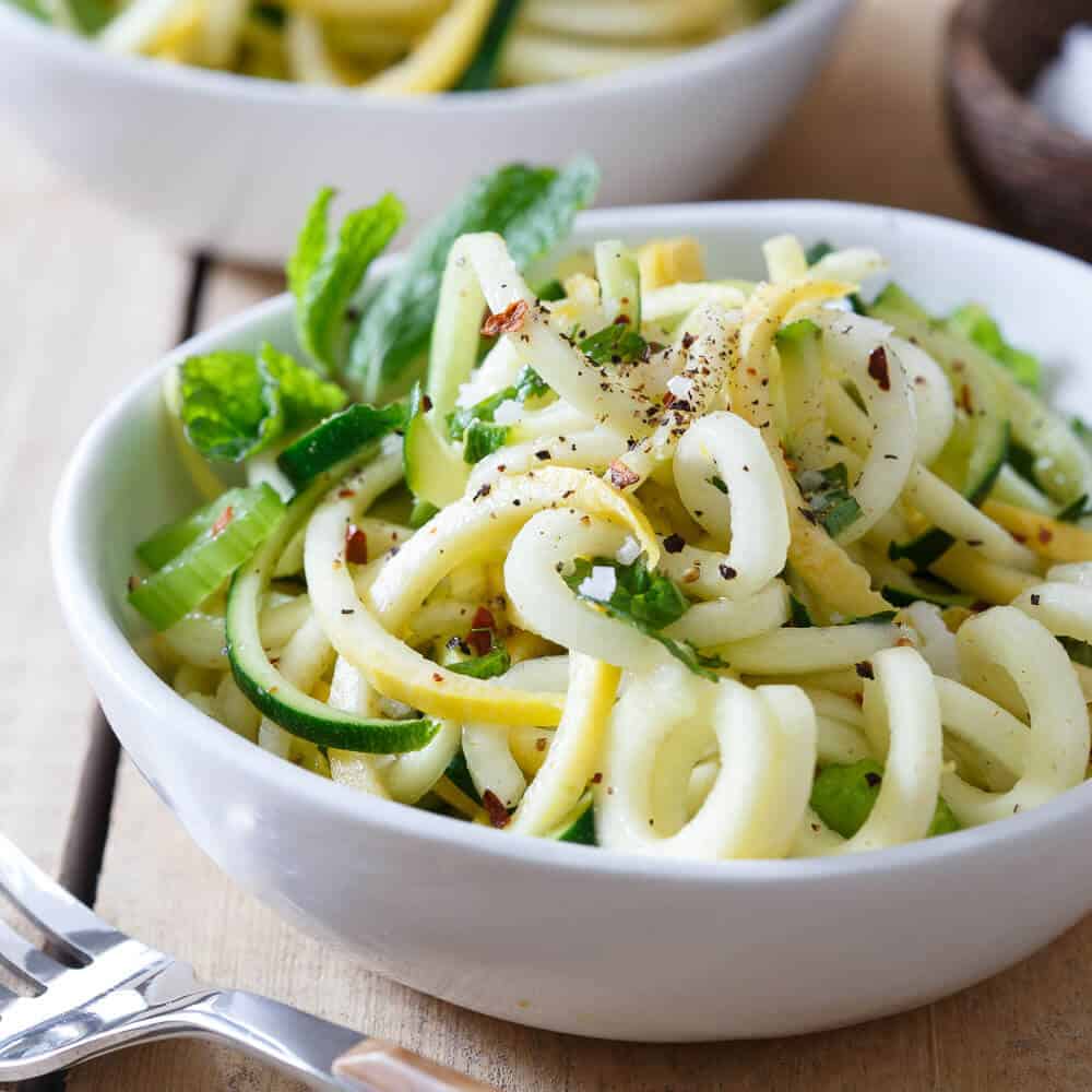 Zucchini and yellow squash noodles in a white bowl with mint garnish.
