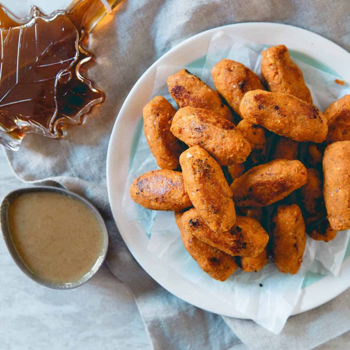 A plate of golden-brown Sweet Potato Tots is on a cloth, with a small bowl of dipping sauce and a maple leaf-shaped container of syrup nearby. The tots are arranged in a pile on parchment paper, ready for dipping and drizzling.