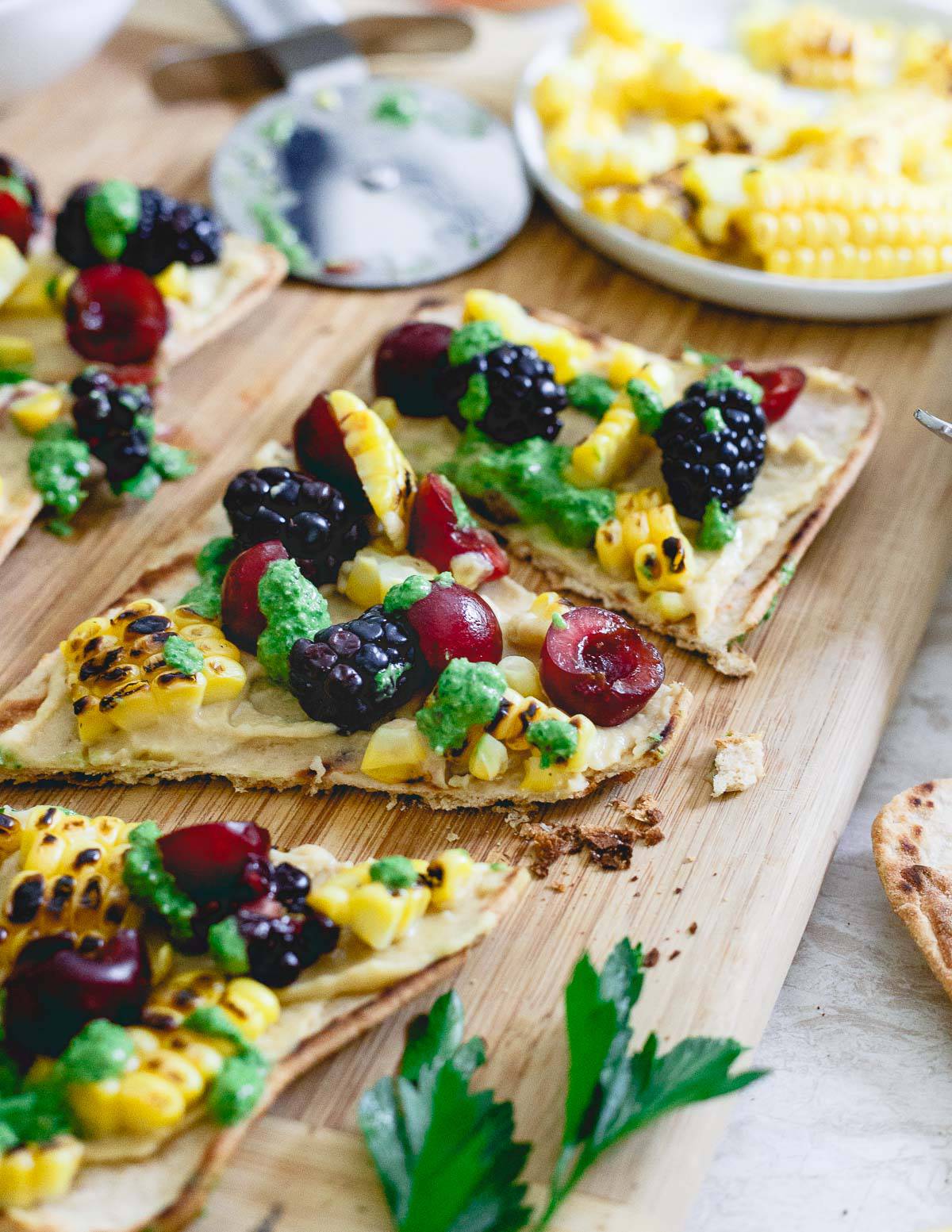 This summer inspired hummus flatbread is topped with grilled corn, berries and drizzled with a fresh roasted garlic parsley pesto. A great pre-dinner bite!