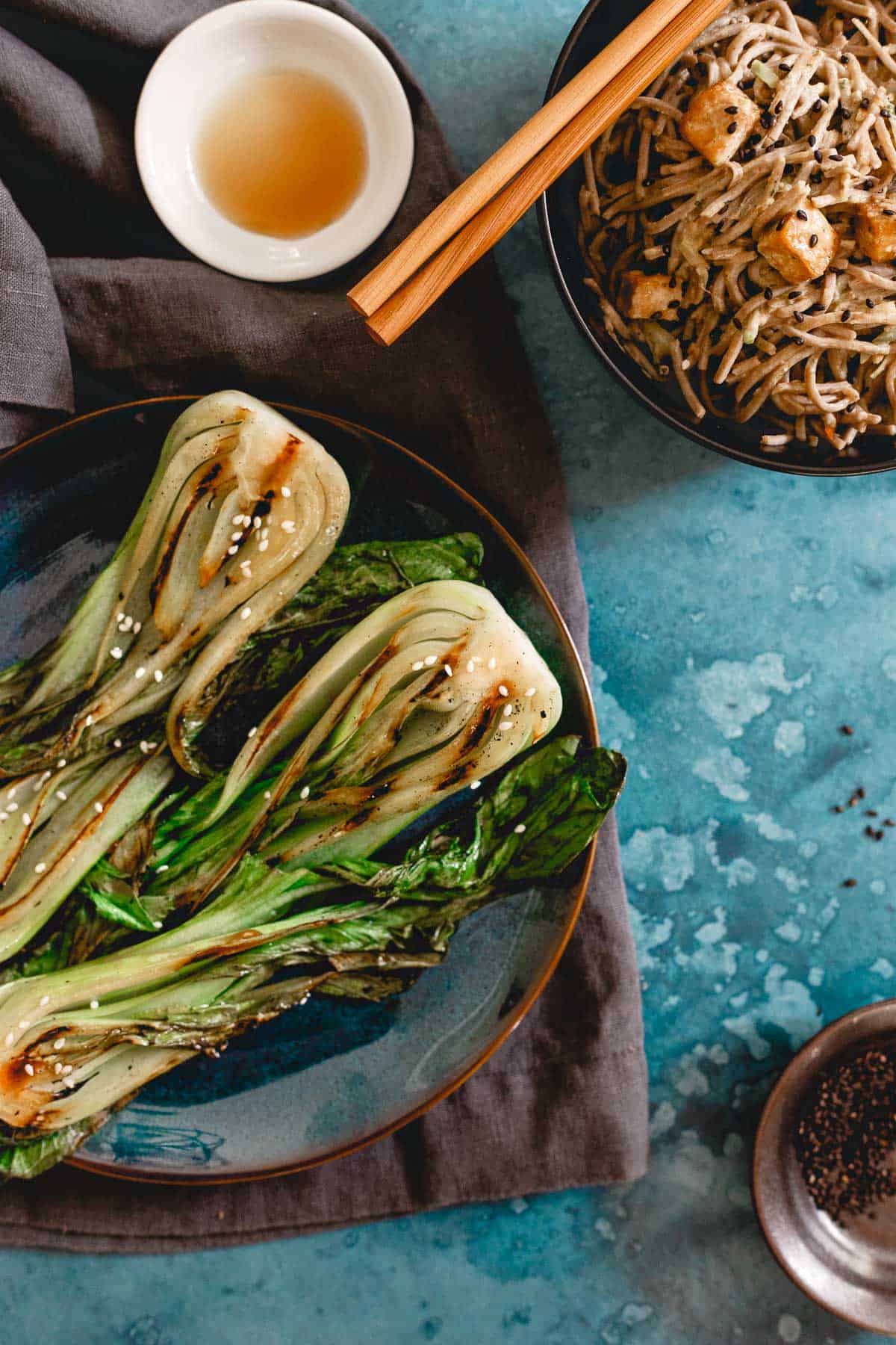 Top view of grilled baby boy choy garnished with sesame seeds next to a bowl of soba noodles.