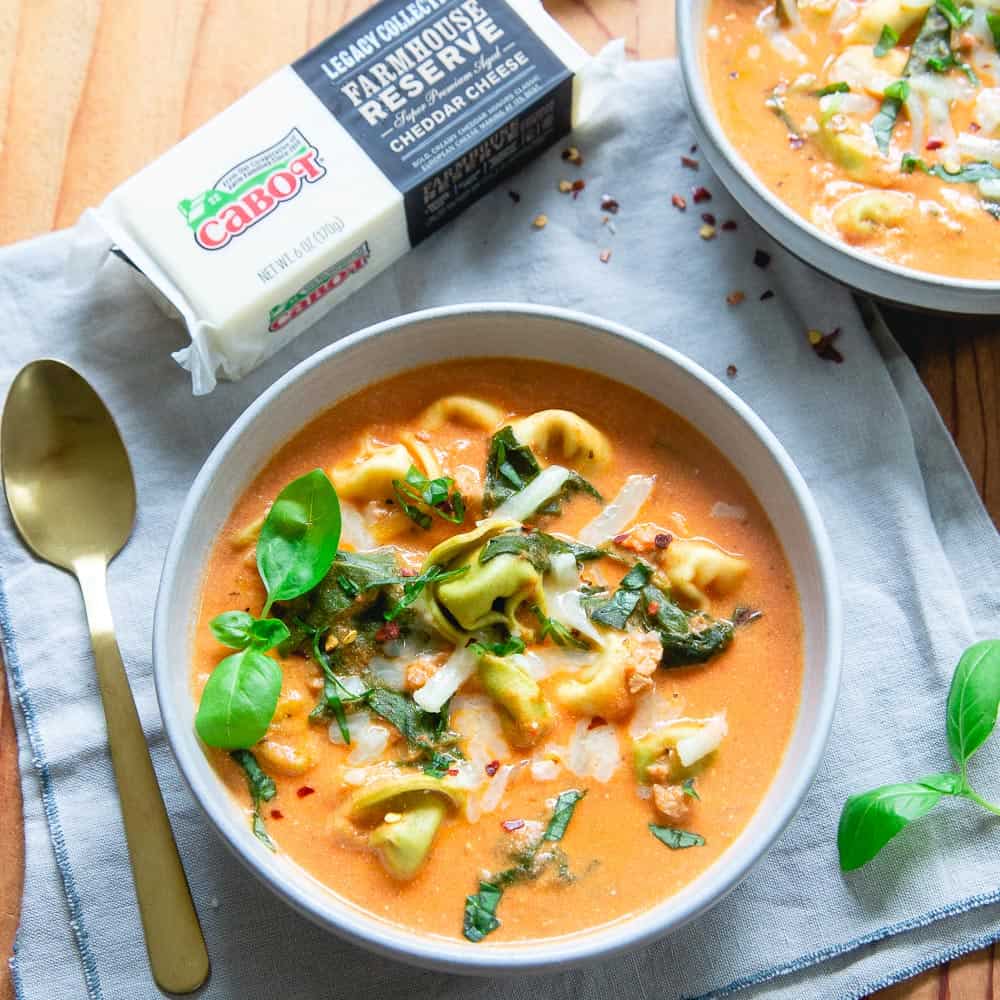 Creamy Instant Pot tortellini soup in a white bowl next to a spoon and a block of cheese.