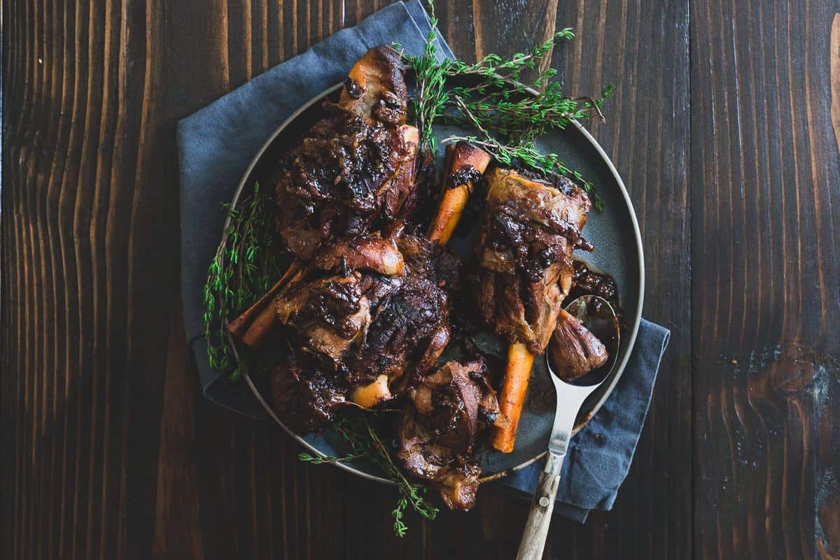 Overhead view of lamb shanks braised in apple cider instead of wine on a dark plate with fresh thyme and a serving spoon.