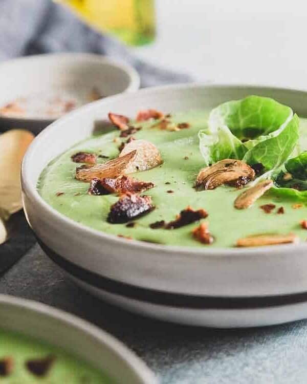 Creamy brussels sprouts soup garnished with roasted garlic chips in a bowl with gold spoons.