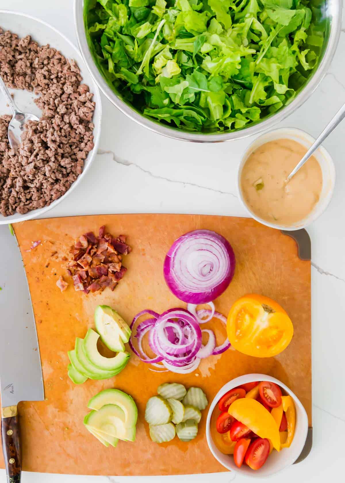 ingredients to make burgers in a bowl including ground beef, prepared lettuce greens, a homemade healthier "special sauce", red onions, tomatoes, bacon, pickles and avocado.