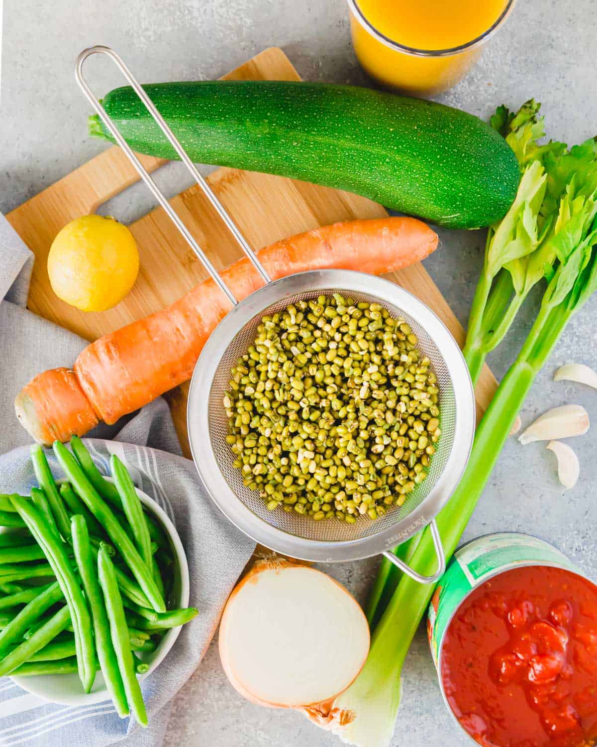 Ingredients to make mung bean soup on a cutting board: rinsed sprouted mung beans, zucchini, carrots, green beans, celery, lemon, crushed tomatoes and vegetable broth.