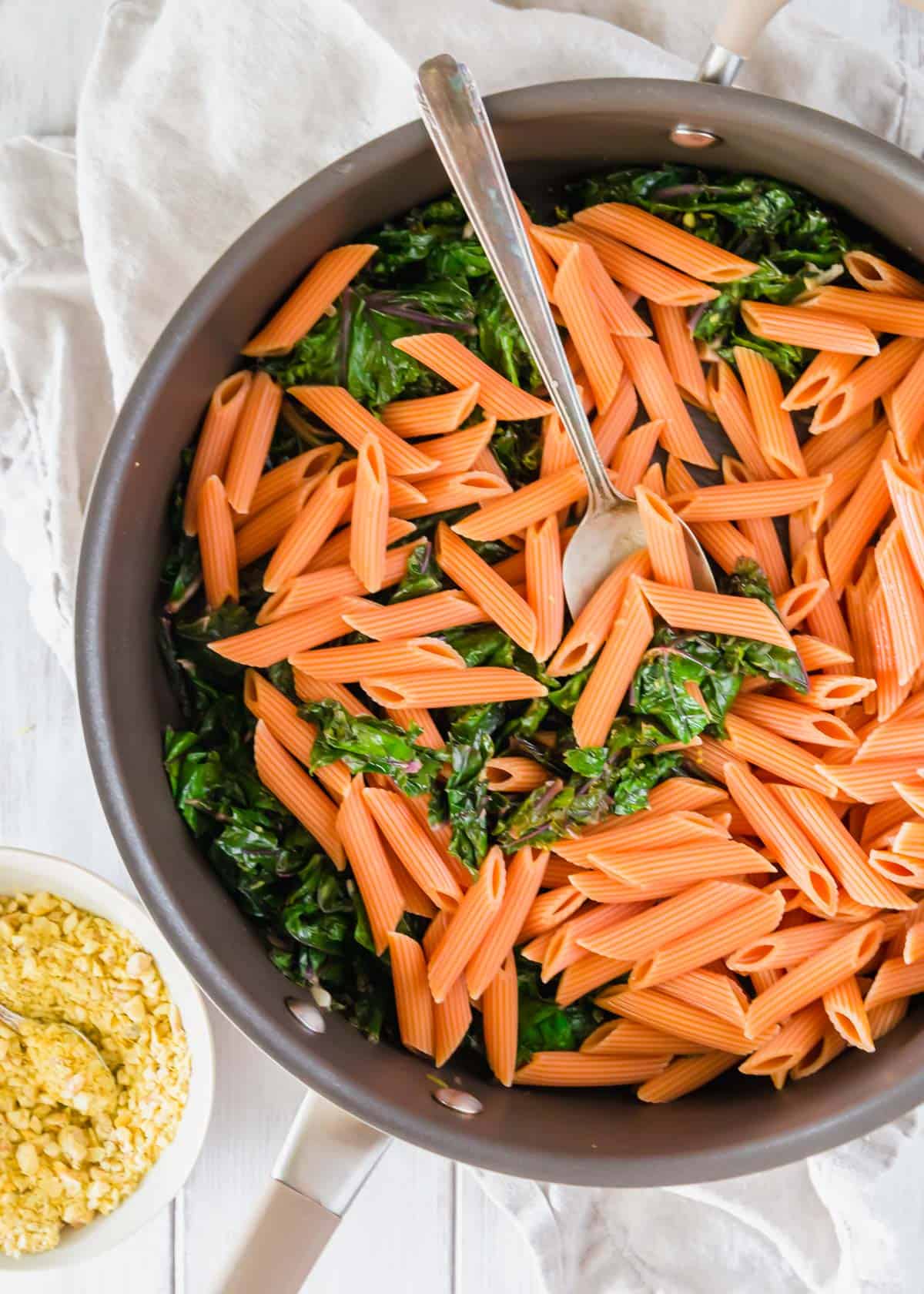 Cooked red lentil pasta tossed with garlic and lemon sautéed kale in a skillet.