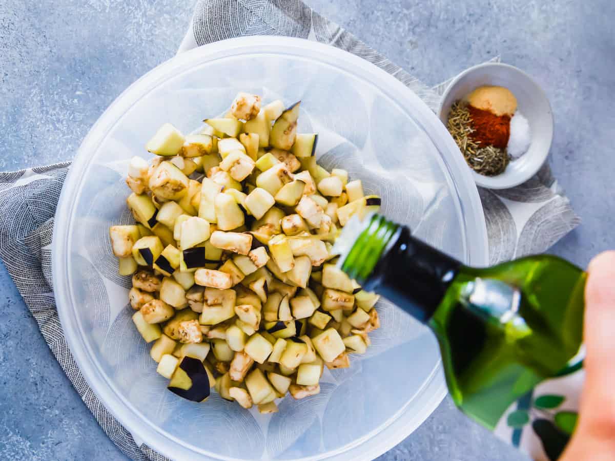 Pouring avocado oil into a bowl with cubed eggplant and spices on the side.