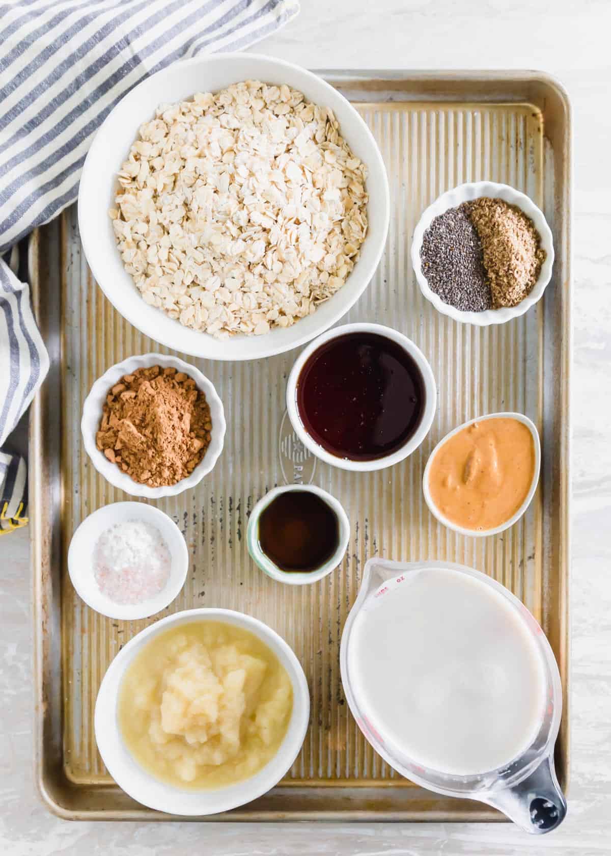 Ingredients to make baked chocolate oats including, rolled oats, chia and flax seeds, maple syrup, cacao powder, almond butter, vanilla, baking powder, salt, applesauce and milk of choice.