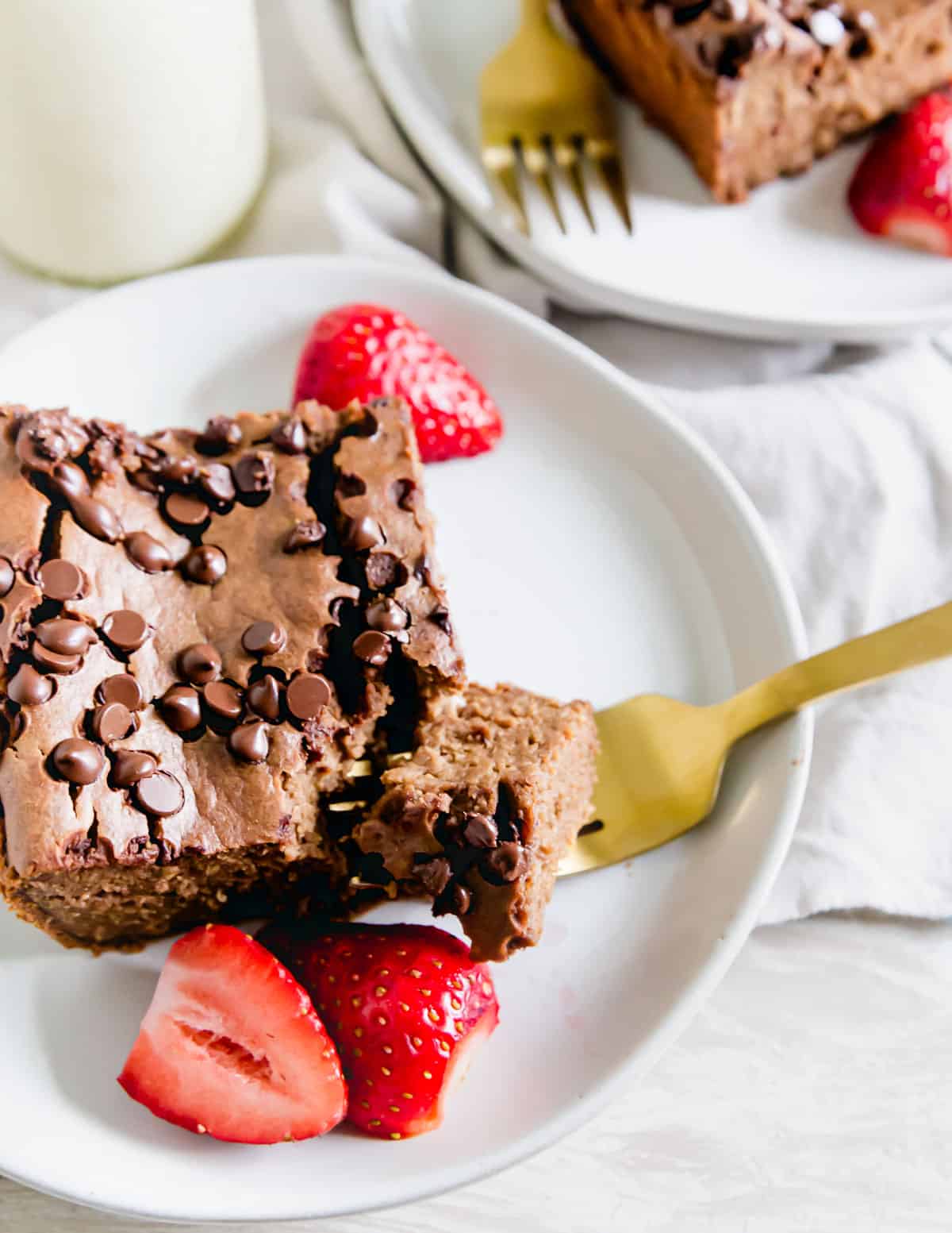 A slice of chocolate baked oats on a plate with a piece on the fork. Fresh strawberries in the background.