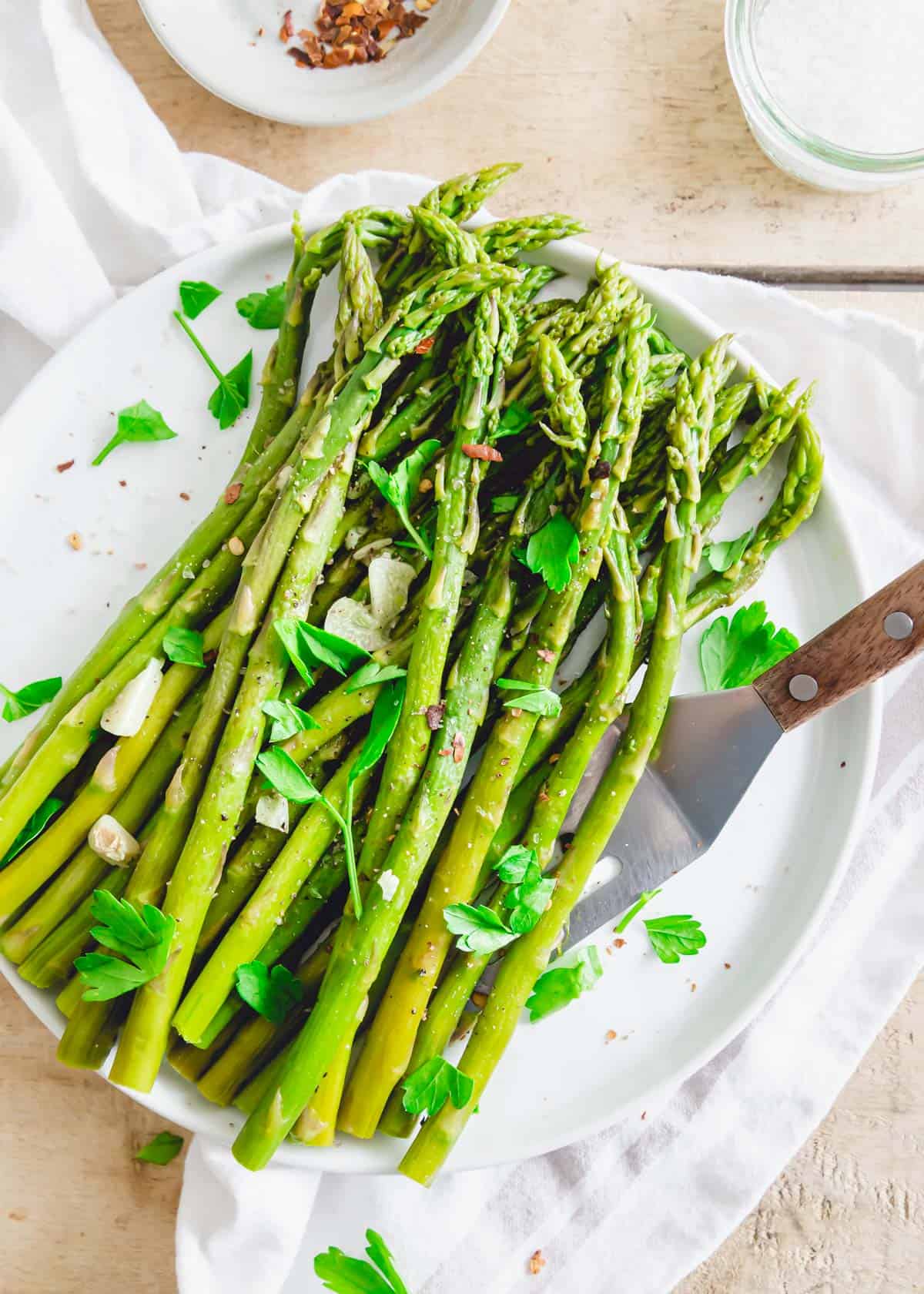 Pressure cooked asparagus on a plate with a serving spatula garnished with garlic, fresh parsley and chili flakes.
