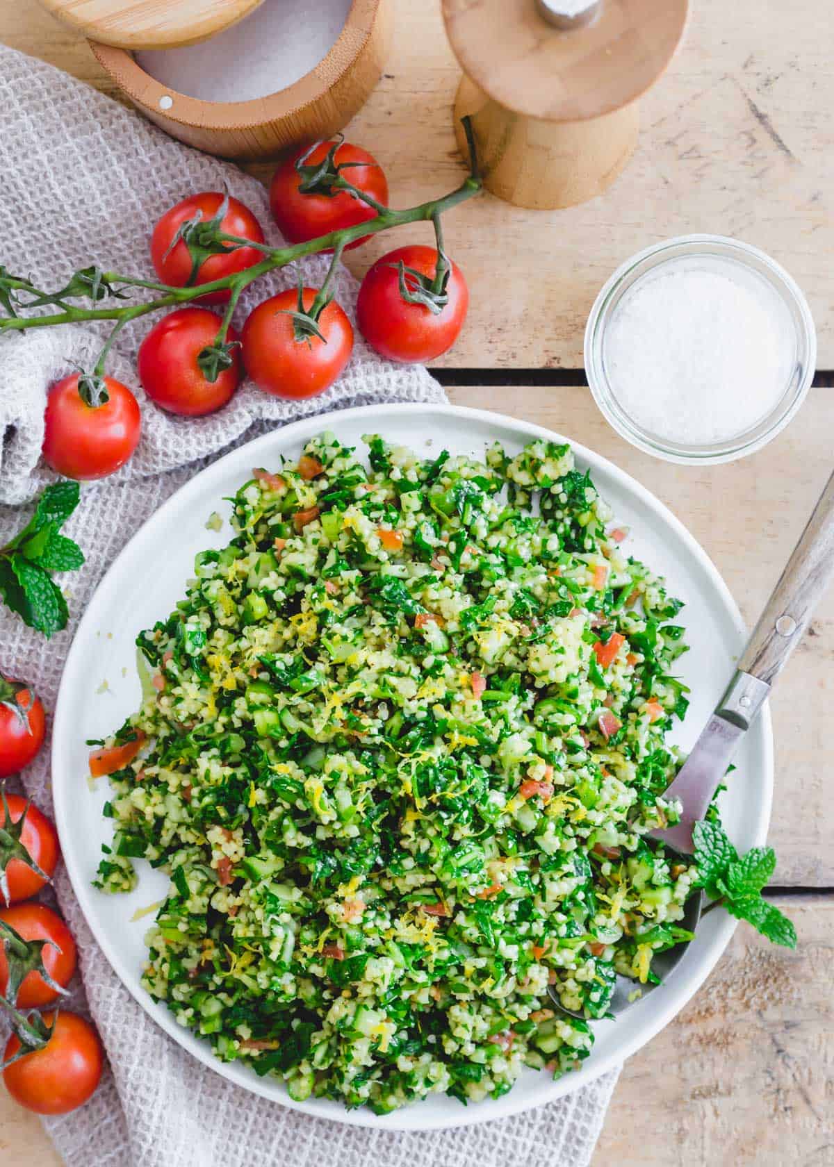 Gluten free tabbouleh made with organic millet on a serving plate with fresh tomatoes, mint, salt and pepper on a table.