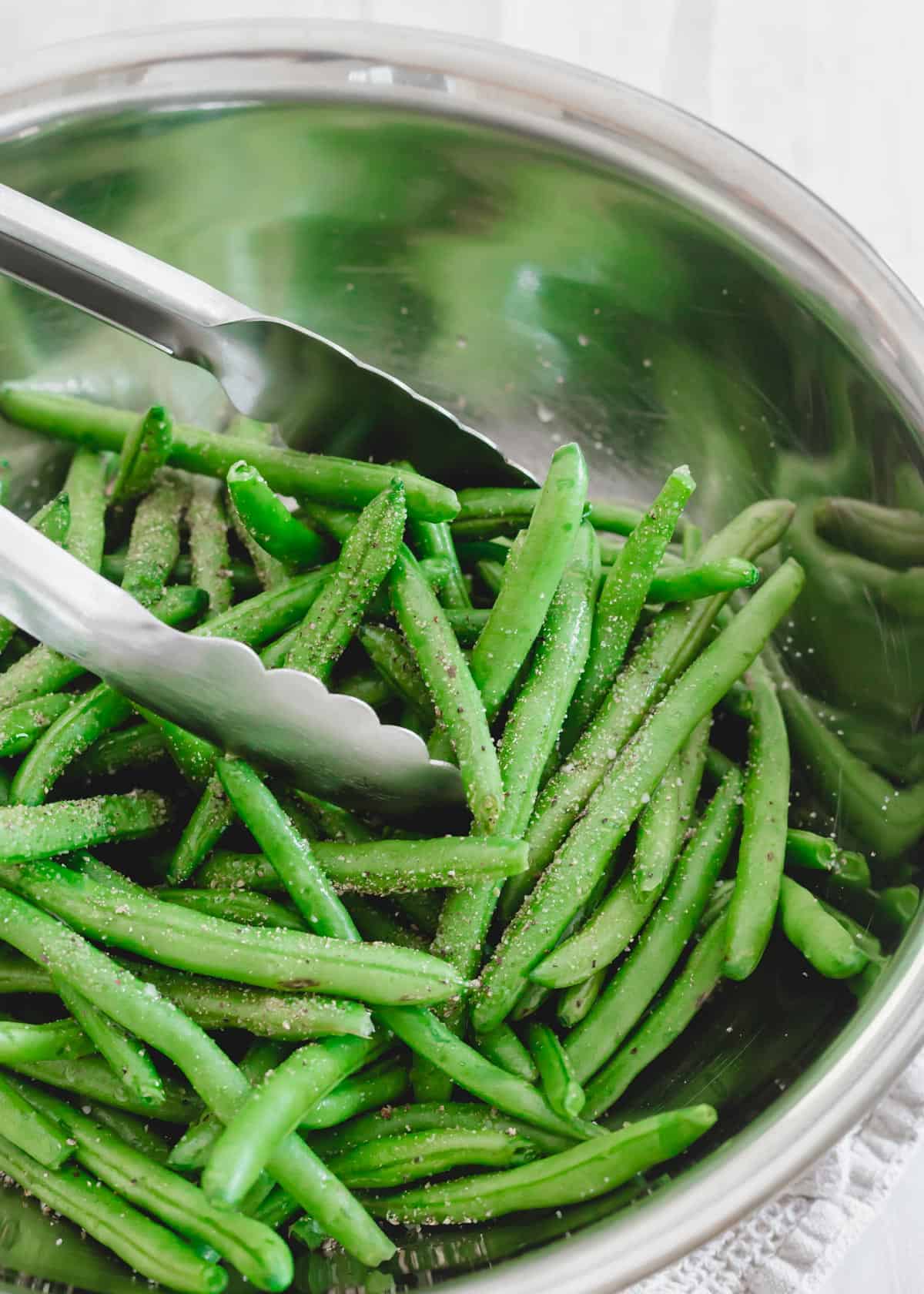 Green beans with garlic powder, salt and pepper in a metal bowl with tongs.