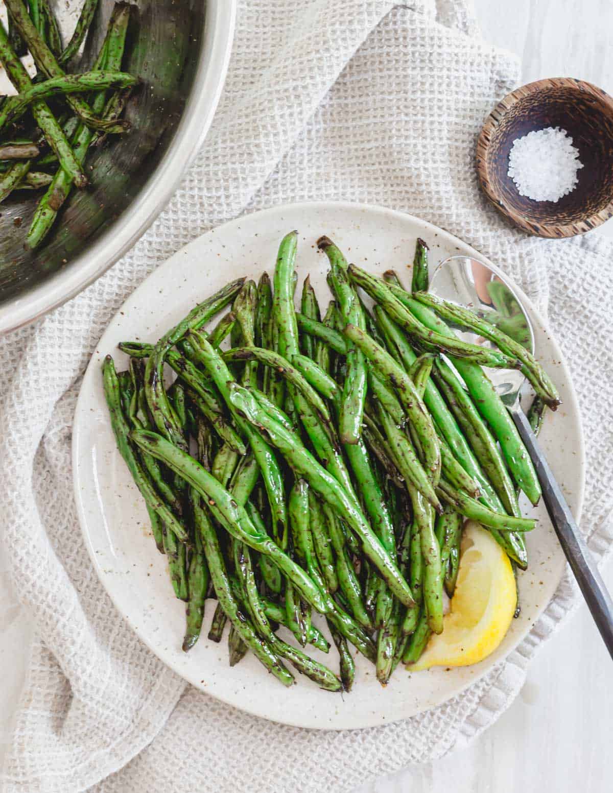 Grilled green beans with garlic and soy sauce on a plate with serving spoon, salt on the side and lemon for serving.