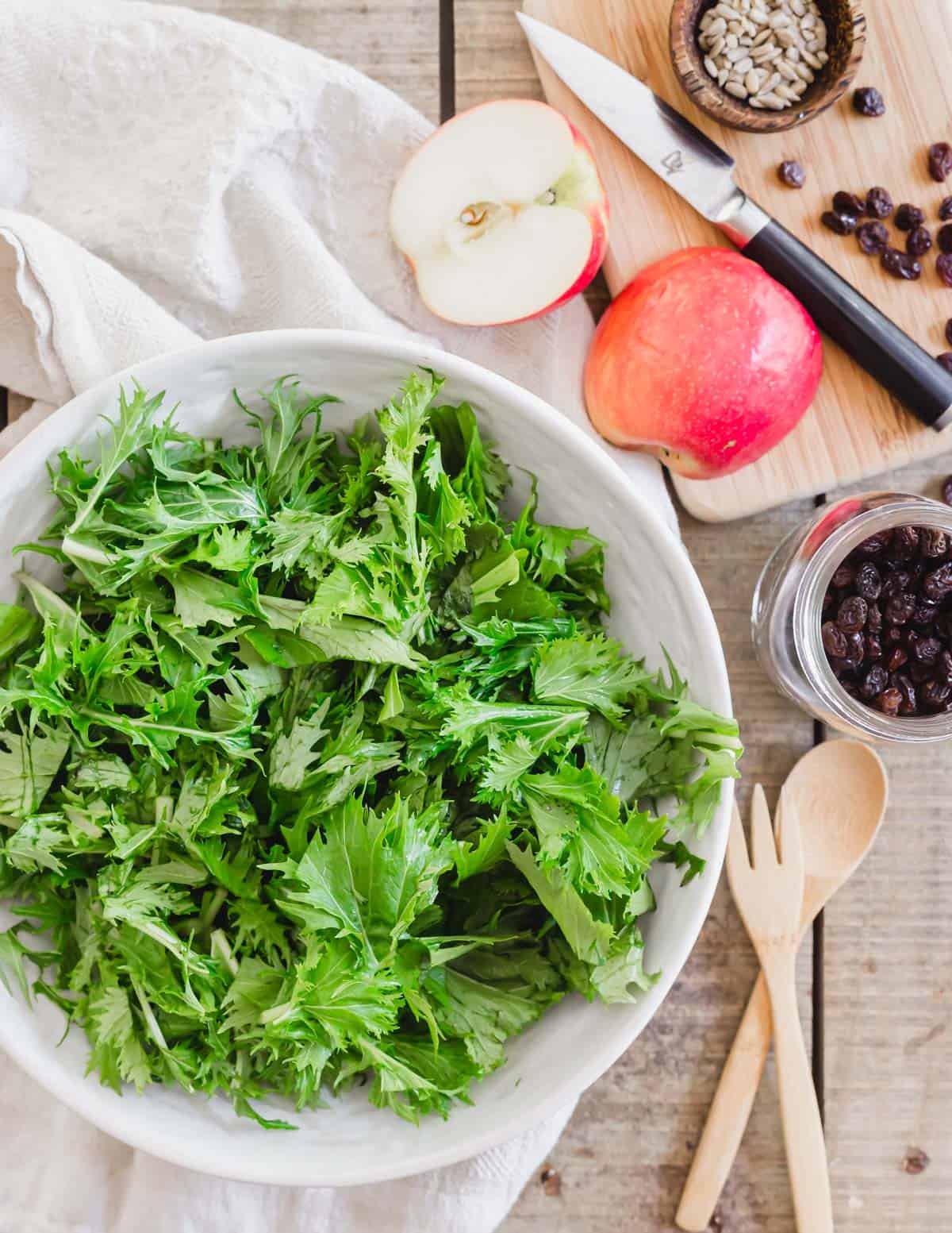 Mizuna lettuce in a bowl with salad components of raisins and an apple on the side.