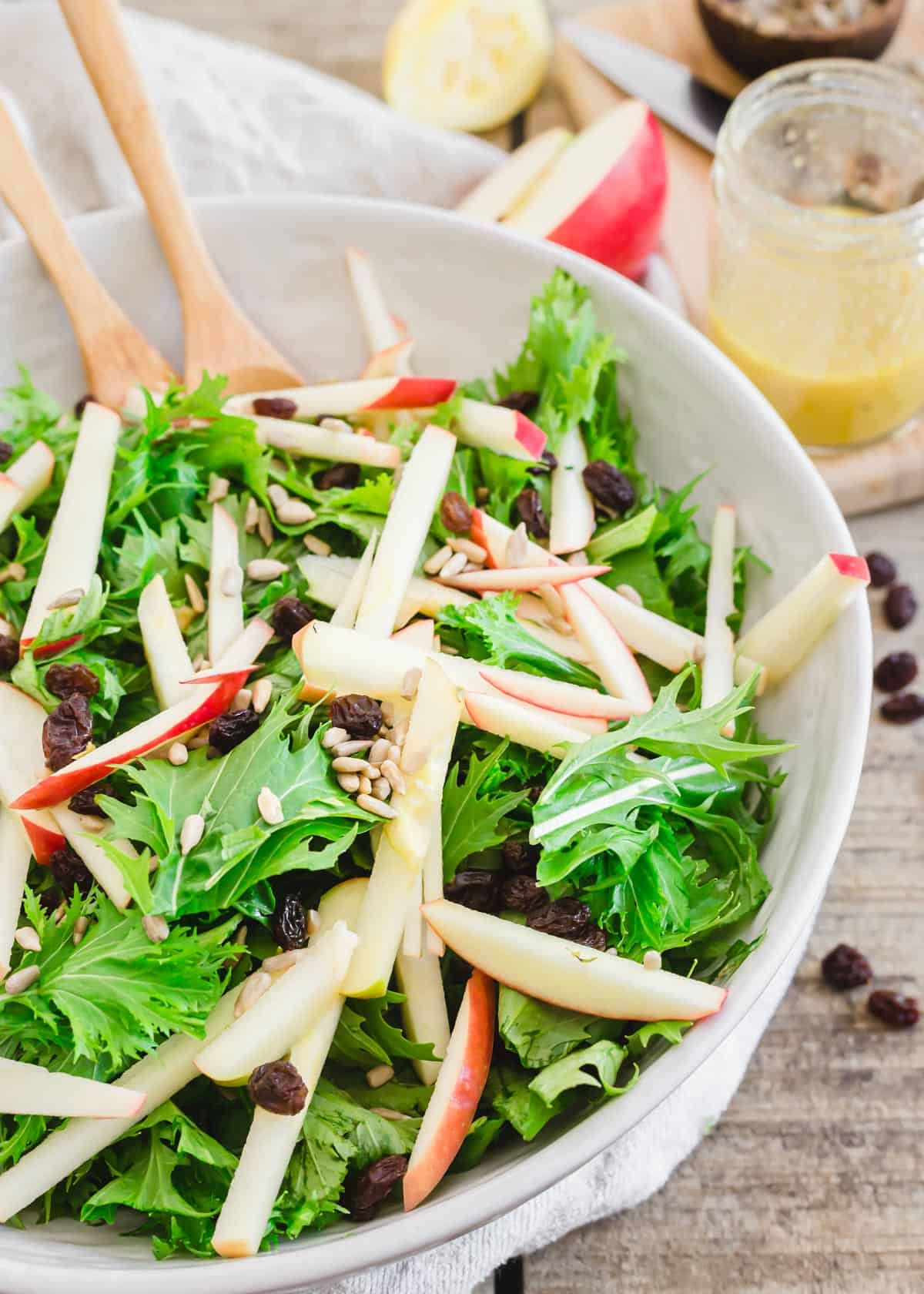 Undressed mizuna salad with apples, sunflower seeds and raisins in a bowl with serving utensils.