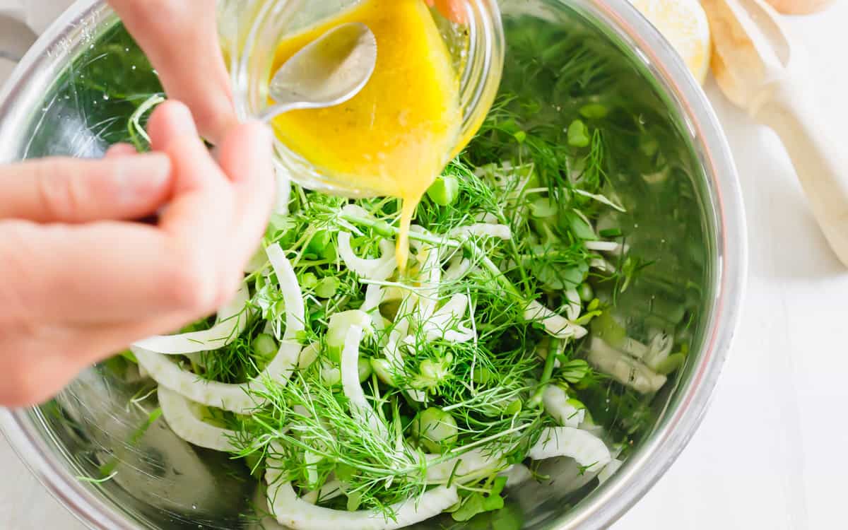 Lemon dijon dressing with honey being poured into a bowl with fresh sliced fennel and baby arugula.