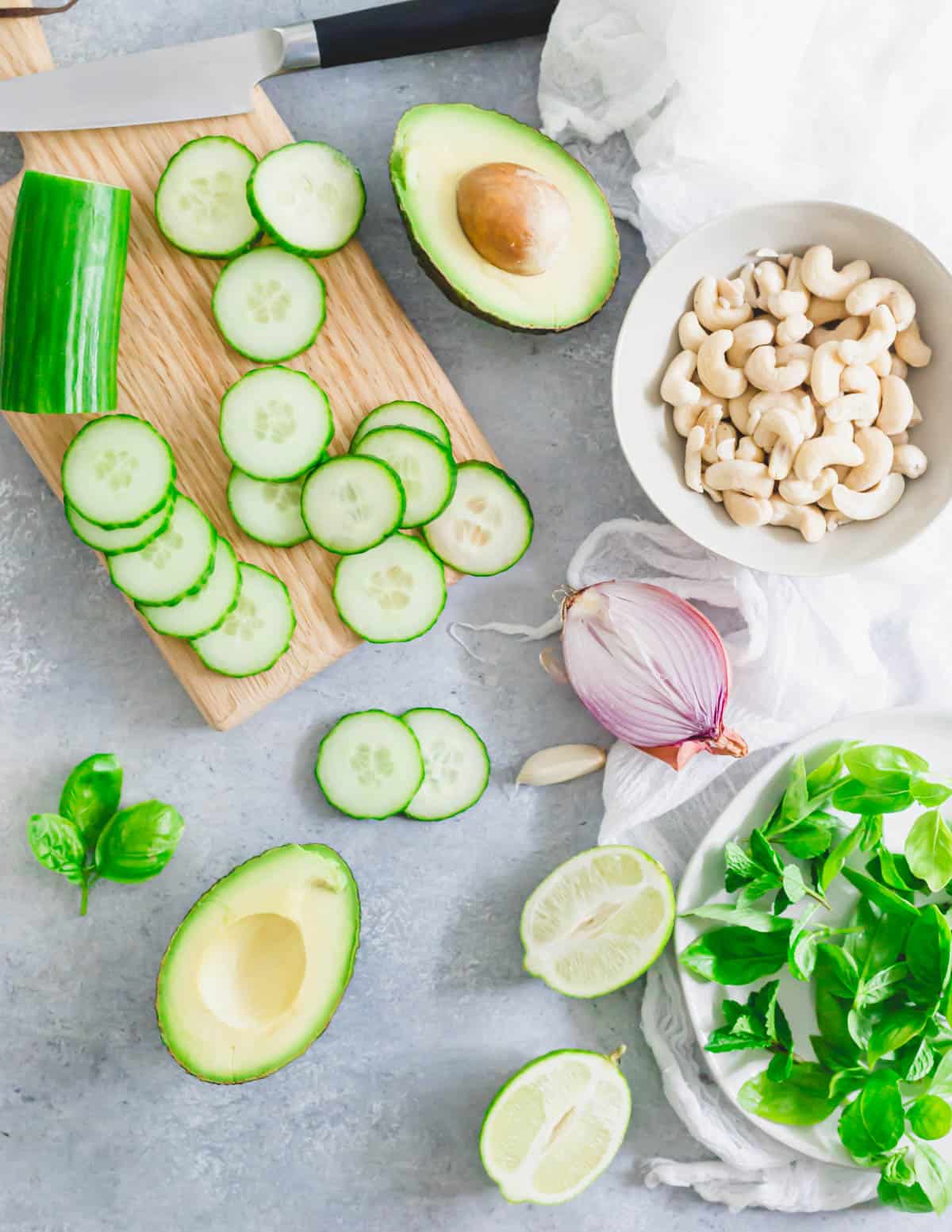 Ingredients to make cucumber gazpacho including cucumbers, soaked cashews, shallot, avocado, mint, basil, lime and garlic.