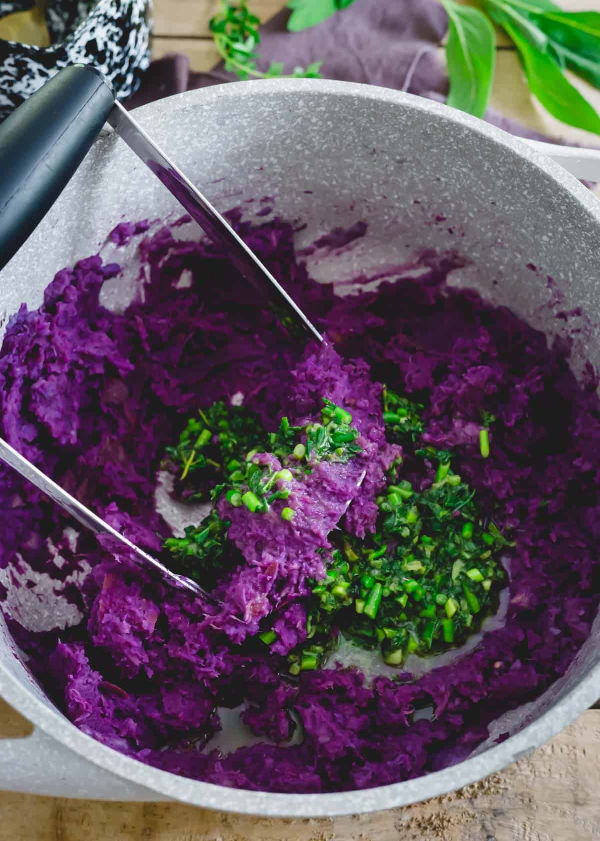Mashed purple sweet potatoes in a pot with a potato masher and herb butter sauce.