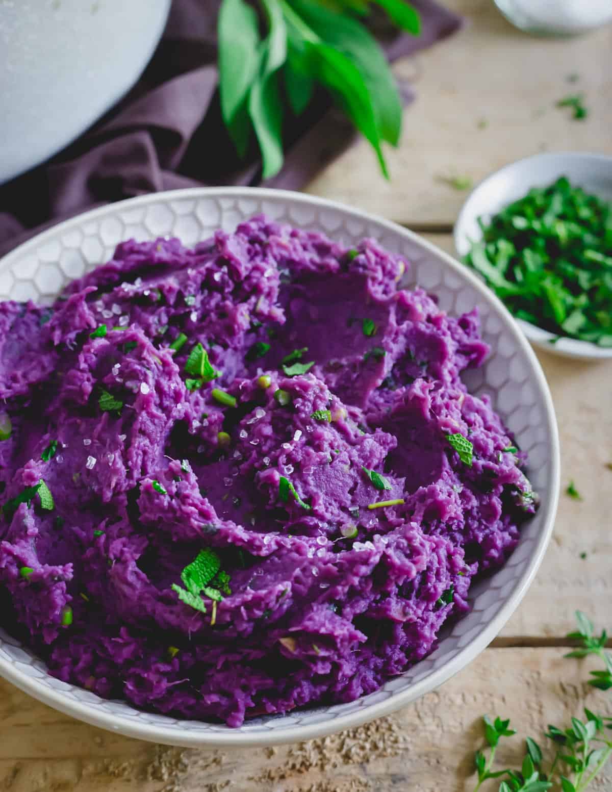 Mashed purple sweet potatoes in a serving bowl with herb butter sauce and flaky sea salt.