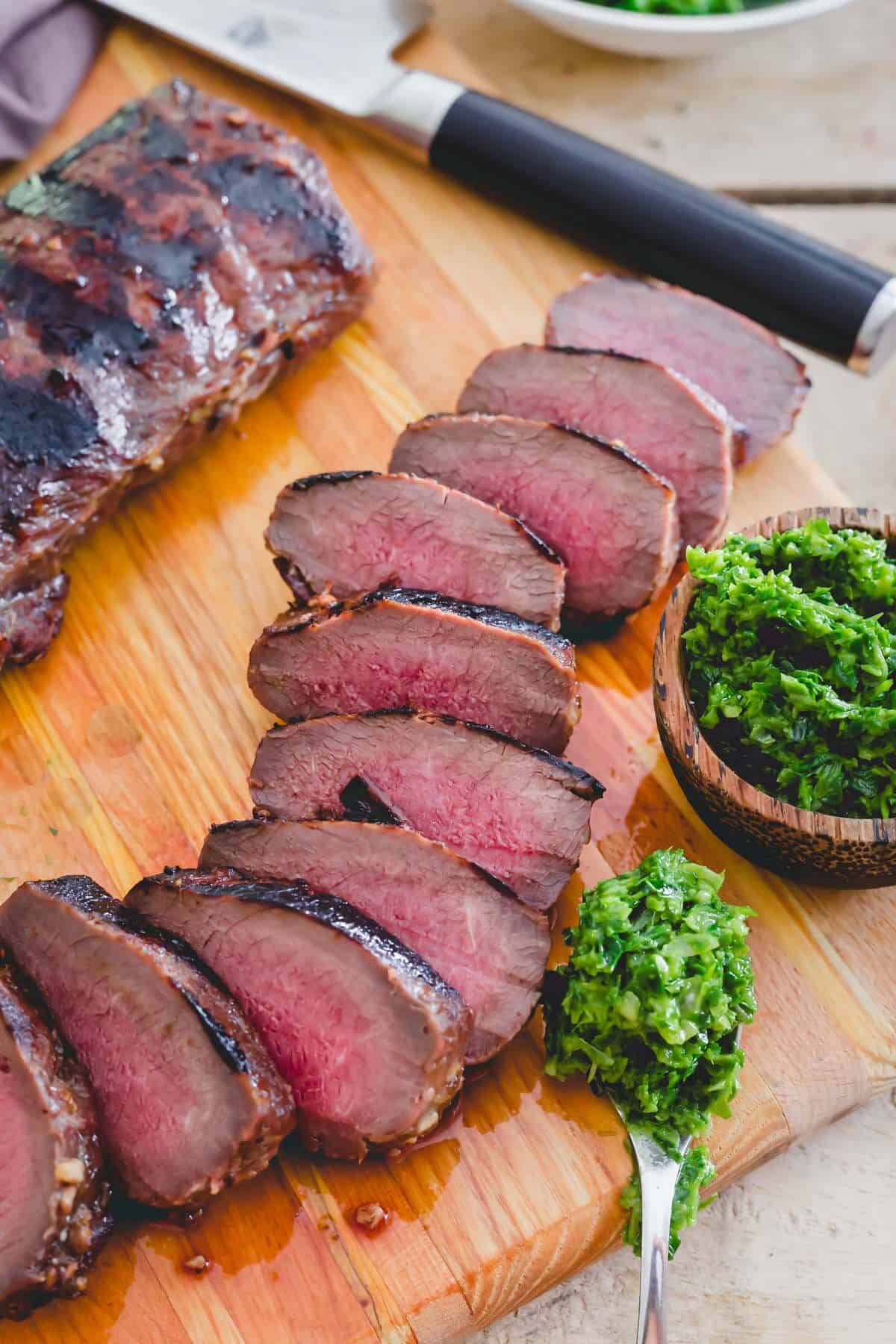 Venison backstrap recipe grilled and sliced on a cutting board with pesto on the side.