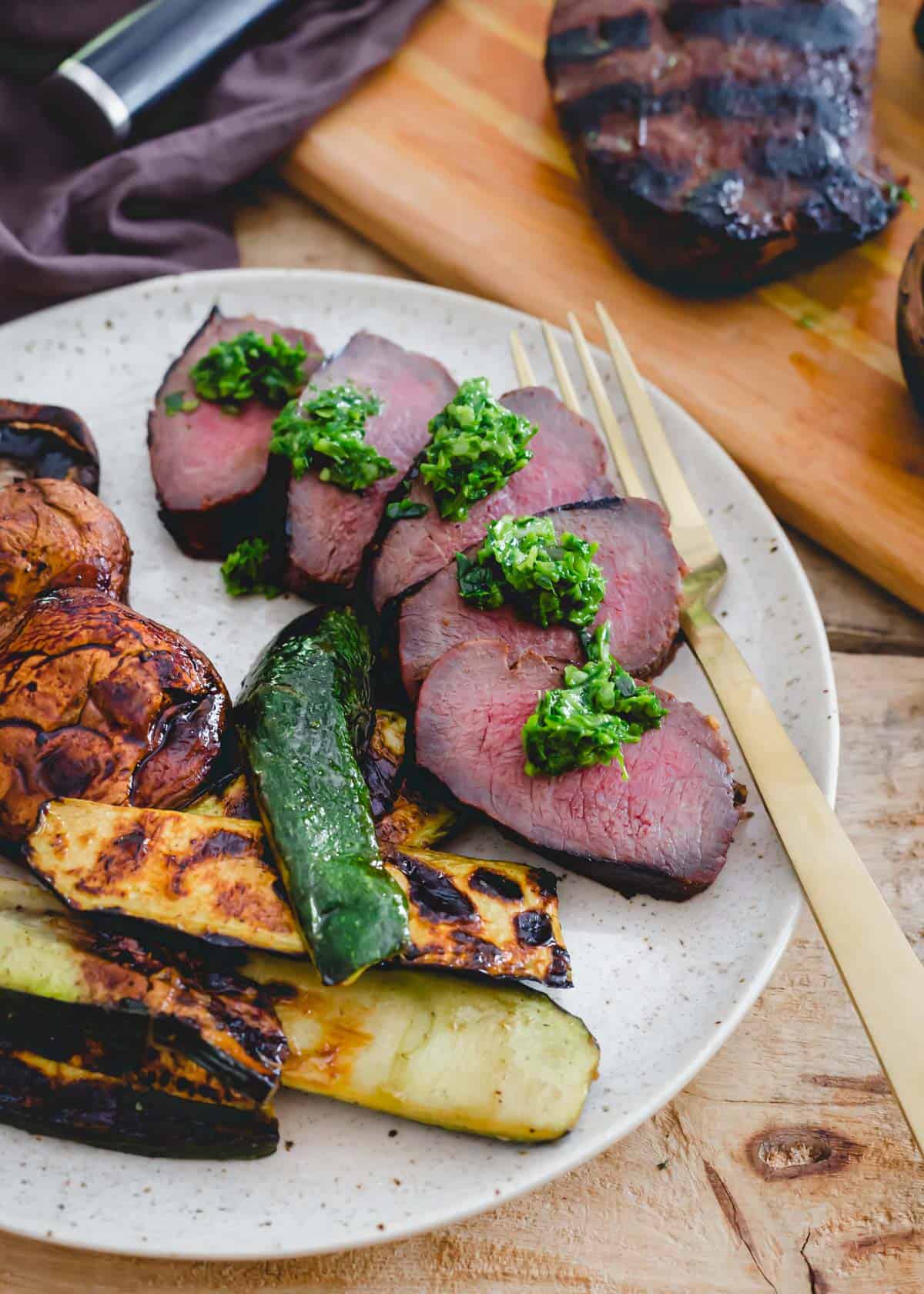 Medium rare grilled venison backstrap sliced and served with pesto and grilled vegetables on a plate.