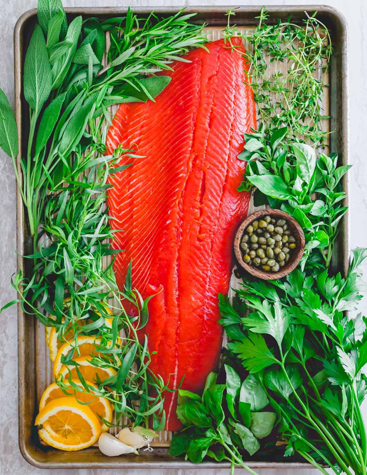 Raw wild sockeye salmon from Alaska with fresh herbs, garlic, capers and orange slices on a baking sheet.