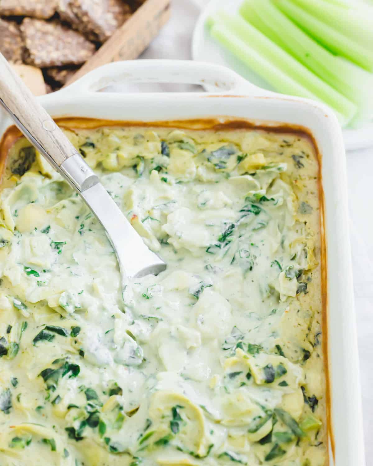 Baking dish of creamy vegan spinach and artichoke dip with a serving spoon.