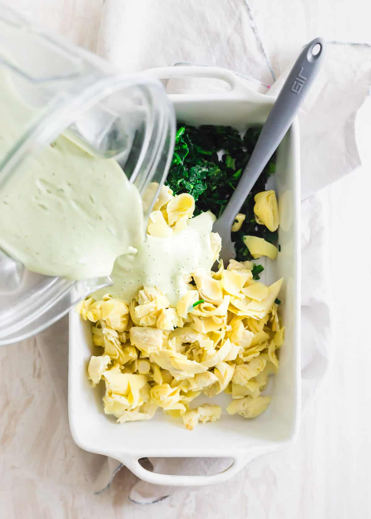 Assembling vegan artichoke spinach dip in baking dish.