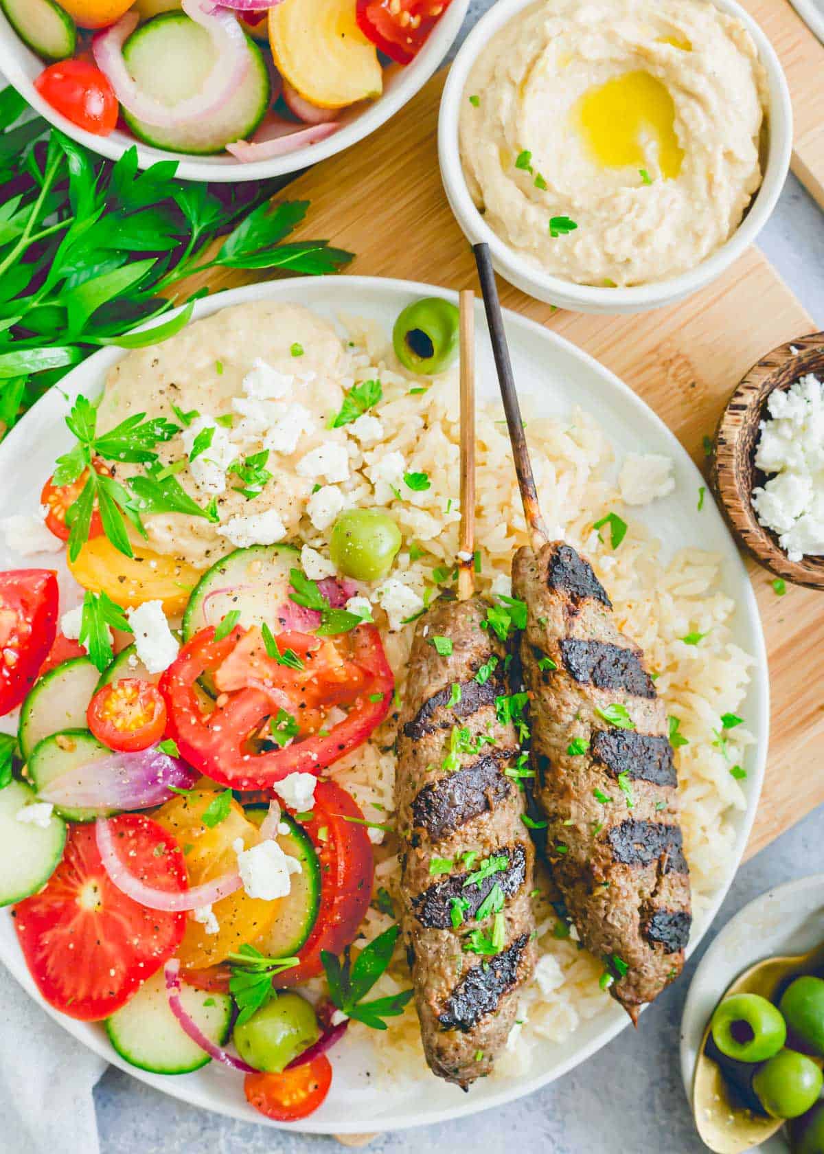 Grilled kafta kabobs made with beef on a plate with rice, tomatoes, cucumbers and red onion and hummus.