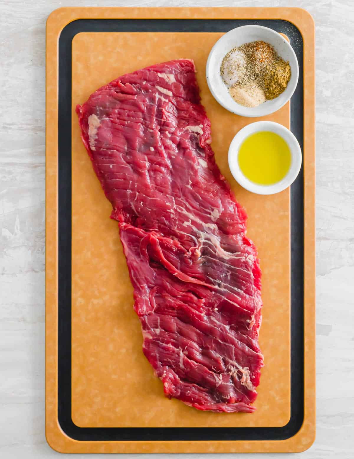 Raw skirt steak on a cutting board with spices and olive oil in bowls to the side.