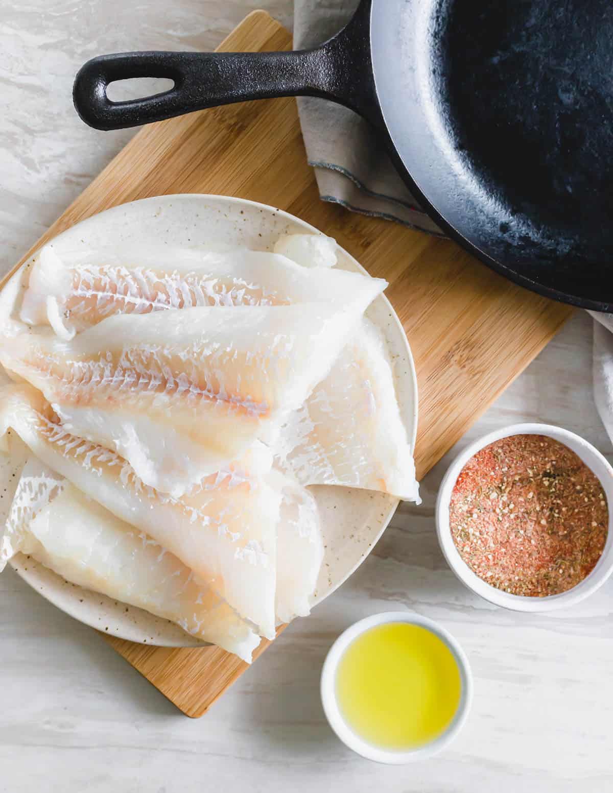 Cod filets on a plate with blackening seasoning and avocado oil next to a cast iron skillet.