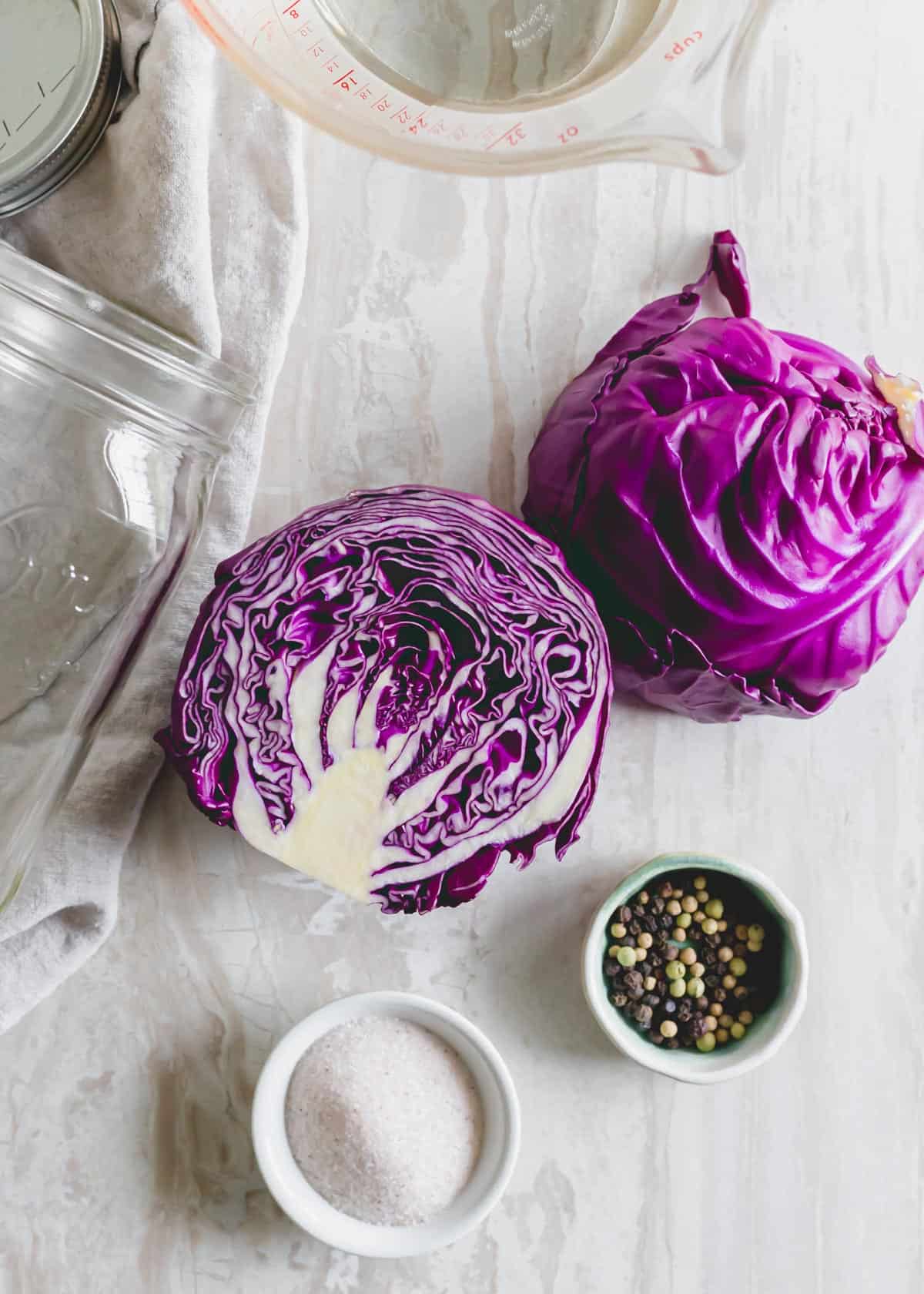 Ingredients to make red cabbage sauerkraut: red cabbage sliced in half, water, salt and peppercorns on a white surface.