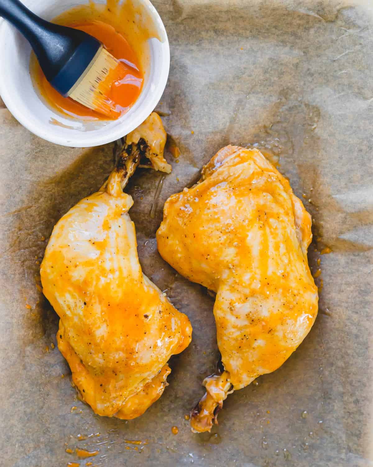 Brushing pressure cooked chicken leg quarters with buffalo sauce on a parchment lined baking sheet.