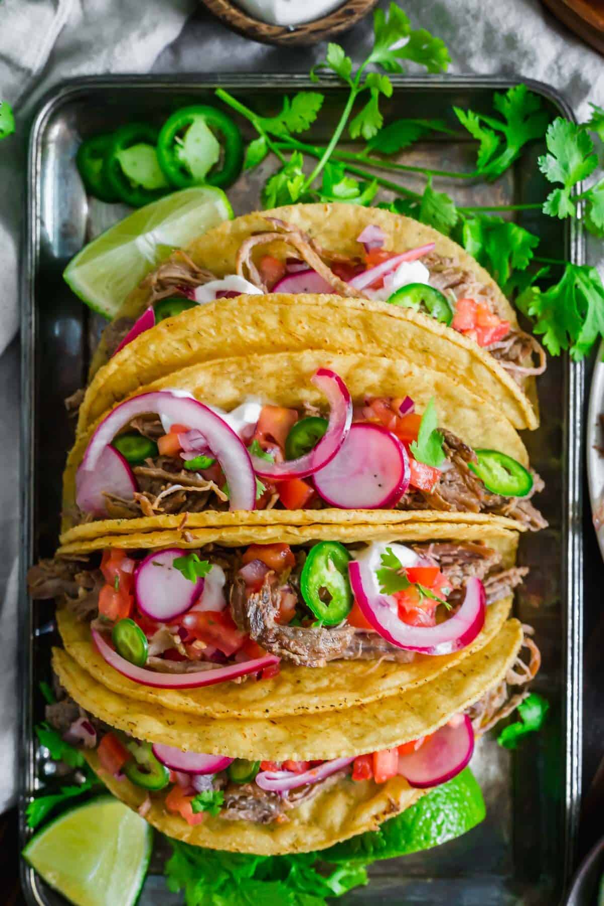 Braised lamb tacos with pickled red onions, radishes, jalapenos, pico de gallo and fresh cilantro on a silver tray.