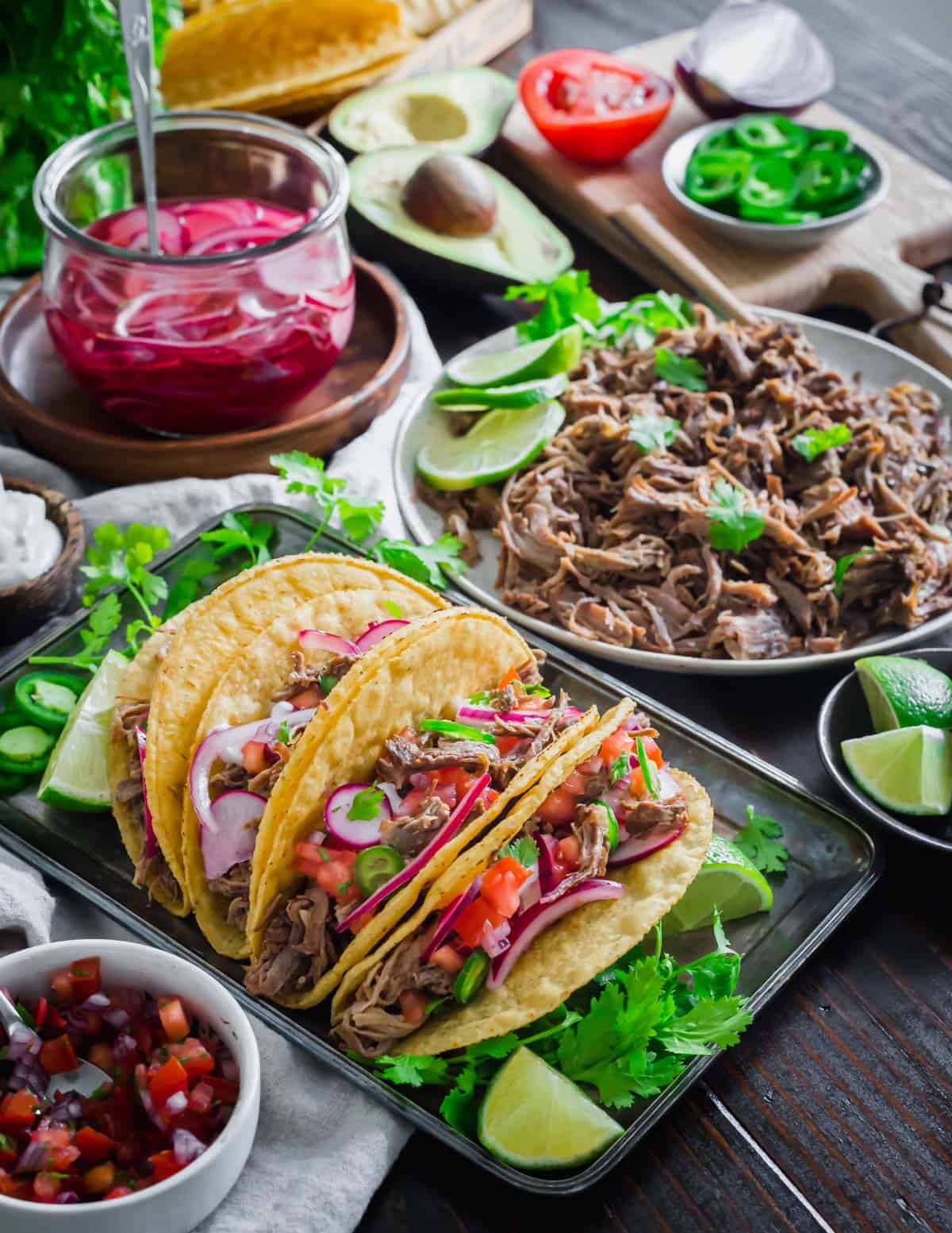Lamb taco spread on a table with lime  wedges, braised lamb meat, pico de gallo, crema, jalapenos, avocado and pickled radishes and red onion.