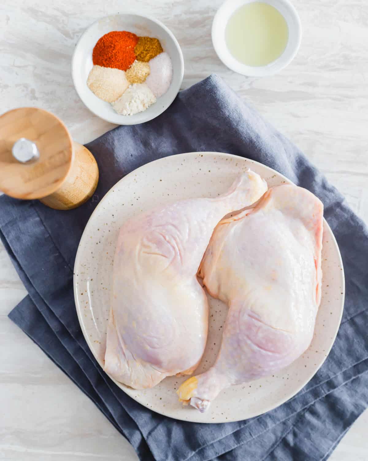 Raw chicken leg quarters on a plate with spice rub and oil to the side.