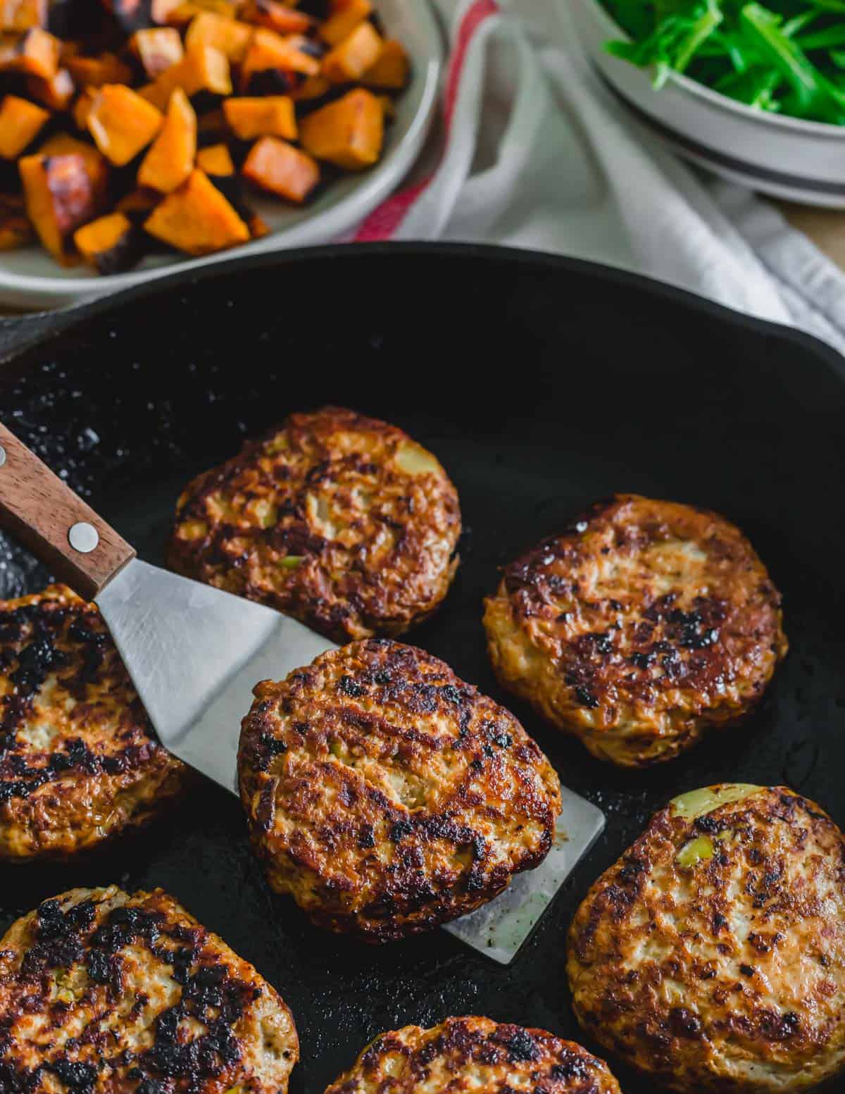 Ground chicken sausage patties in a cast iron skillet with a metal spatula.