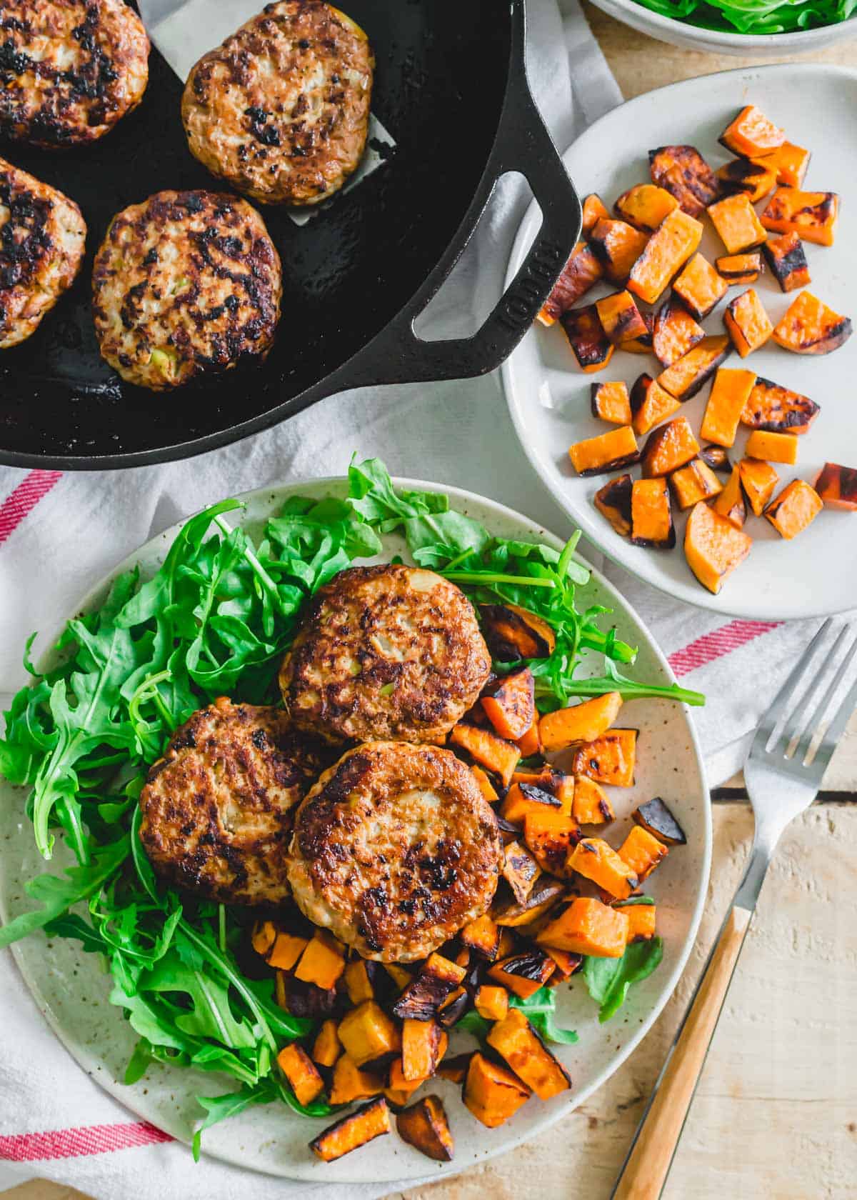 Chicken breakfast sausage patties with apples and onions over a bed a arugula and crispy sweet potatoes on a plate.