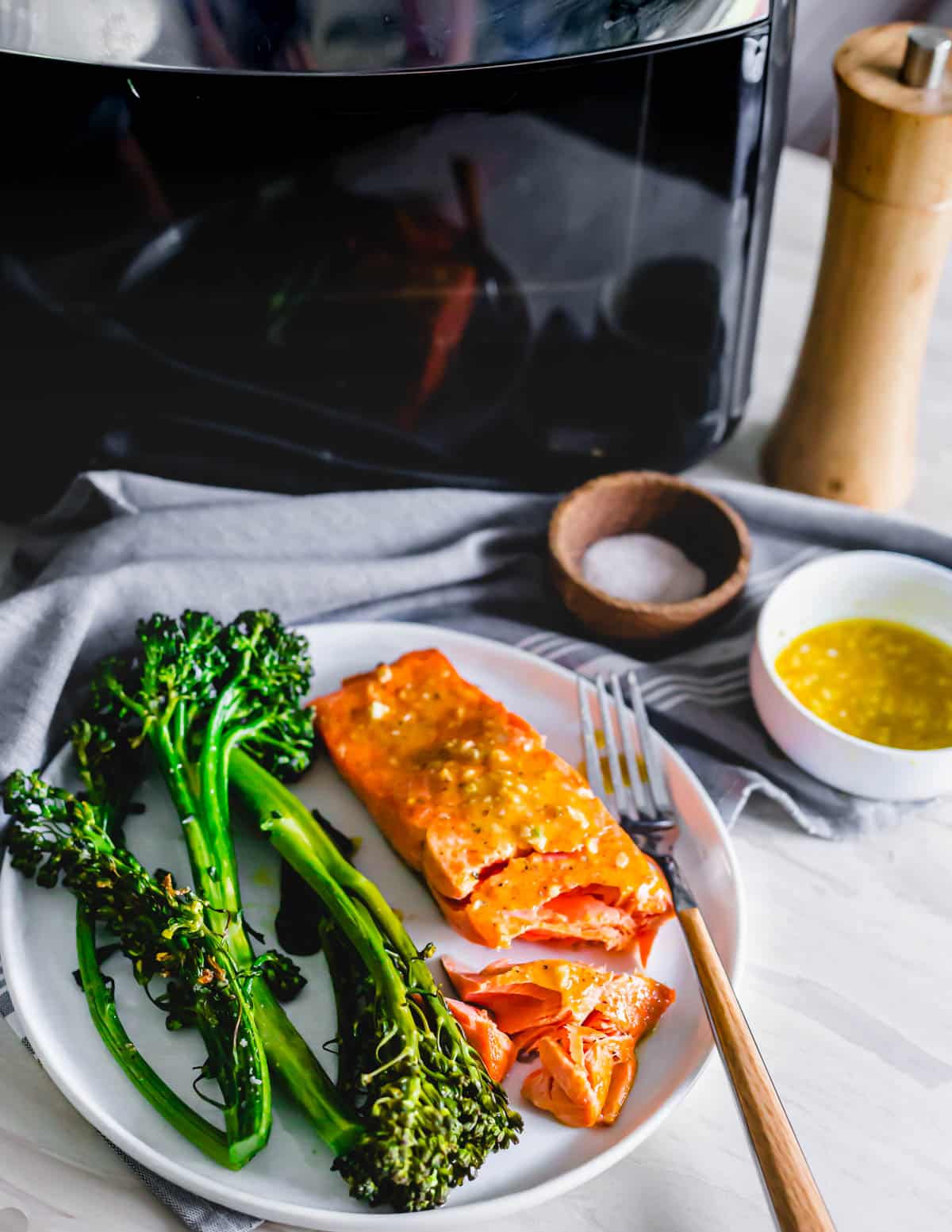 Frozen salmon filet cooked in a Pampered Chef air fryer on a plate with broccolini and maple mustard sauce on the side.