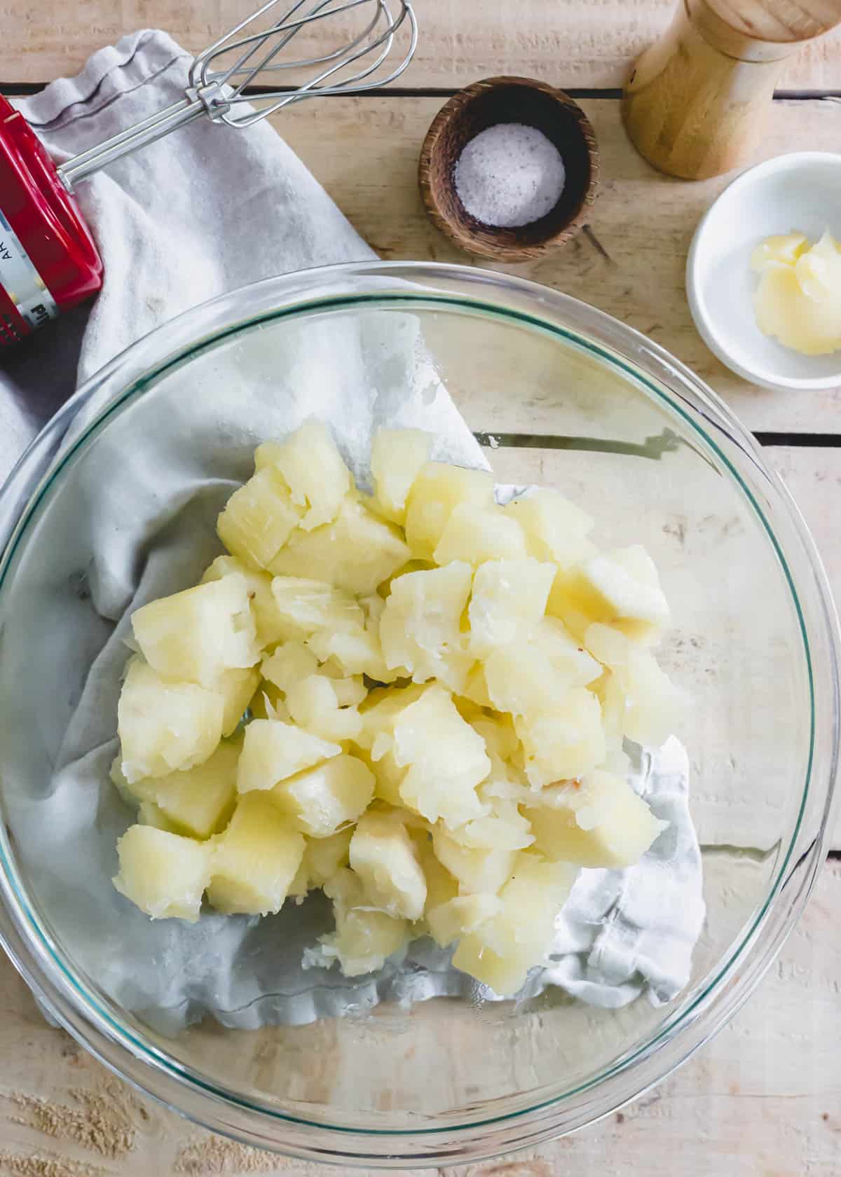 Boiled yucca root in a glass bowl with salt, pepper, ghee and a handheld mixer on the side.