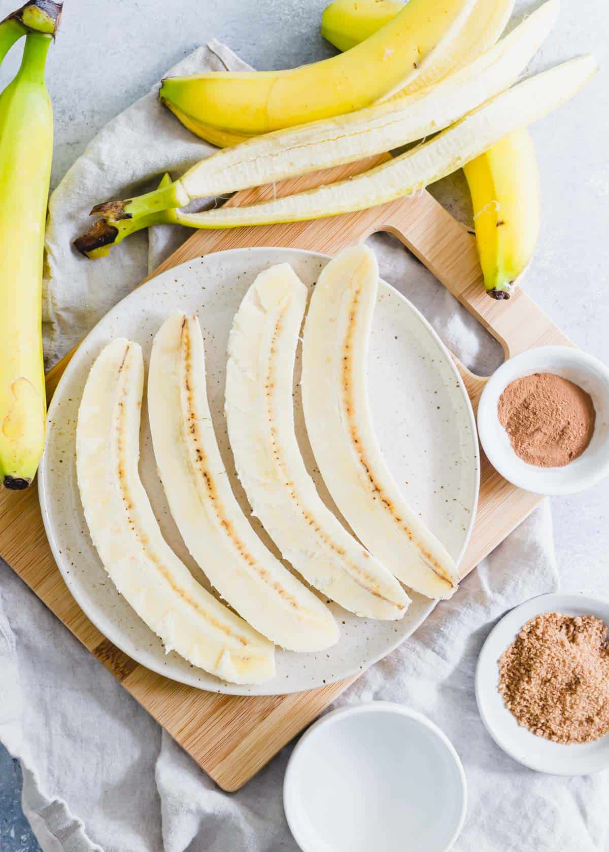 Bananas halves on a plate with cinnamon spices, brown sugar and melted coconut oil in bowls on the side.